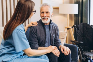 Bakıcı, gülümseyen yaşlı bir adama yardım ediyor ev ziyareti sırasında destek ve şefkat sunuyor. Arka planda tekerlekli sandalye