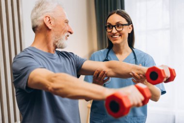 Kıdemli adam küçük dambıllarla rehabilitasyon egzersizi yapıyor, gülümseyen kadın bir hemşireden rehberlik alıyor.