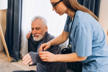 Hemşire yaşlı bir adama süveterini giymesi için yardım ediyor, evde günlük yaşam aktiviteleri için destek ve yardım teklif ediyor.