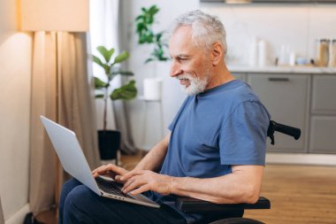 Özürlü, gülümseyen, tekerlekli sandalyede otururken dizüstü bilgisayarda yazan, evdeki teknolojiyle bağlantı kuran son sınıf öğrencisi.