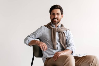 Modern gündelik kıyafetler giyen, güler yüzlü, sakallı Latin adam oturmuş kameraya bakıyor. Beyaz arka planda yalnız çalışan yakışıklı bir iş adamı. İş konsepti