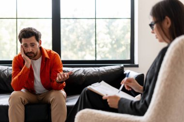 Stresli orta yaşlı adam terapi seansı sırasında sorunları tartışıyor. Erkek hasta kadın doktorla konuşuyor, modern ofiste birlikte otururken notlar alıyor. İletişim konsepti