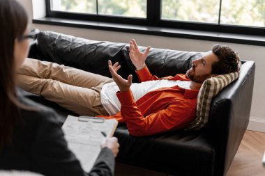 Stresli, üzgün bir adam terapi seansı sırasında modern ofiste rahat bir koltukta uzanıp terapistle konuşuyor. Dinleyen ve not alan kadın doktor. Terapi konsepti