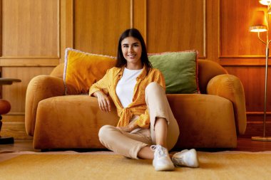 Oturma odasında rahatça kameraya bakarak gülümseyen güzel bir kadın. Evde poz veren mutlu bir kadın model. Reklam kavramı