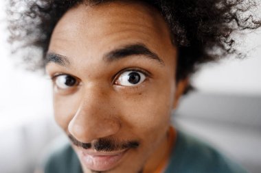 Kıvırcık saçlı, bıyıklı, açık gözlü genç bir adam kameraya tuhaf ve şaşkın bir ifadeyle bakıyor.