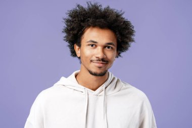 Afro-Amerikalı genç adam gülümsüyor. Mor arka planda beyaz bir kapüşonluyla poz veriyor.