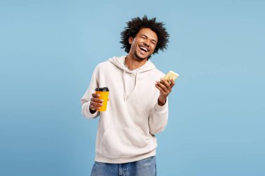 Genç Afro-Amerikan bir adam güçlü bir şekilde gülüyor, elinde bir telefon ve kahve fincanı var, mavi bir arka planda duruyor.