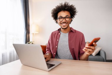 Kameraya gülümseyen Afro-Amerikalı adam elinde akıllı bir telefonla uzaktan çalışıyor ve dizüstü bilgisayar kullanıyor.