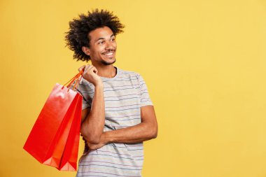 Gülümseyen genç adam elinde kırmızı alışveriş torbalarıyla parlak sarı bir arka plana bakıyor. Perakende, tüketim ve satış temsilcisi