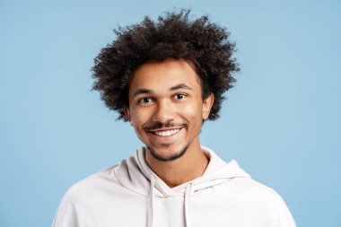 Genç Afro-Amerikan adam mutlu bir şekilde gülümsüyor, olumlu bir ifadeye sahip, mavi bir stüdyonun arka planındaki kameraya bakıyor.