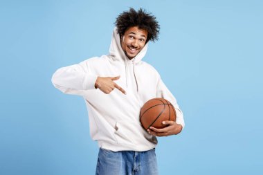 Gülümseyen, elinde basketbol topu tutan ve mavi arka plandaki kameraya bakan mutlu genç siyahi adam.