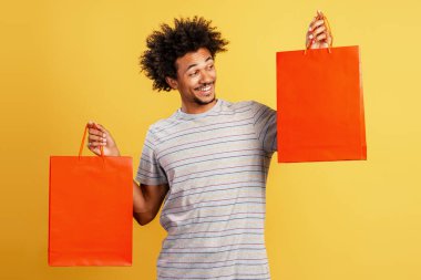 İki canlı turuncu alışveriş torbası tutan mutlu Afrikalı genç adam, tüketim ve perakende terapisinin keyfini çıkarıyor.