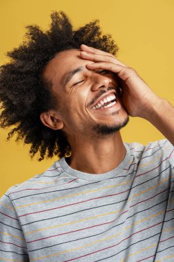 Afro saçlı genç adam gülüyor, elleriyle yüzünü örtüyor, sarı arka planda neşe ve olumlu duygular gösteriyor.