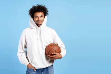 Kapüşonlu Afro-Amerikalı bir adam elinde basketbol topuyla mavi arka planda kameraya gülümsüyor.