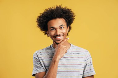 Mutlu genç siyah adam gülümsüyor, kameraya bakıyor, sarı arka planda parmakları çenesinde olan bir hareket düşünüyor.