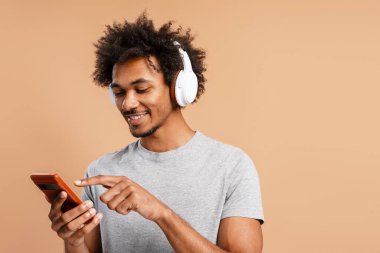 Genç adam gülümsüyor, beyaz kulaklık takıyor, müzik dinliyor ve açık kahverengi arka planda akıllı bir telefonla iletişim kuruyor.