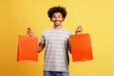 Sarı arka plandaki kameraya gülümseyen iki boş turuncu alışveriş çantası tutan mutlu genç adam.