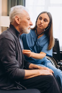 Evde steteskopla yaşlı adamın kalbini dinleyen güzel bir hemşire. Modern klinikte oturan olgun bir hasta. Sağlık hizmeti kavramı
