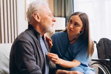 Mavi üniformalı güzel hemşire sağlık ziyareti sırasında steteskop kullanarak olgun bir adamın kalbini ya da ciğerlerini kontrol ediyor. Sağlık hizmeti kavramı