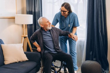 Gülümseyen, güzel bir bakıcı, tekerlekli sandalyedeki yaşlı adama yardım ediyor. Mutlu doktor, modern oturma odasında olgun bir hasta yetiştiren fizyoterapist. Rehabilitasyon kavramı