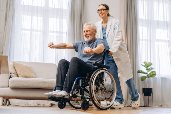 Elderly man doing physiotherapy exercises with caregiver at home. Senior patient sitting in wheelchair in living room. Rehabilitation concept