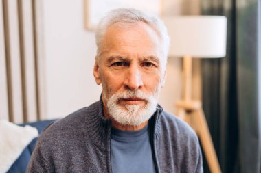 Gri sakallı ve bıyıklı yaşlı bir adam, sıradan bir kazak giyiyor, kameraya ciddi bir ifadeyle bakıyor, bilgelik ve deneyim tasvir ediyor.