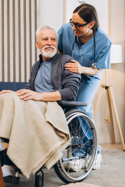 Smiling, happy woman, nurse supporting senior man in wheelchair at home. Beautiful doctor supporting male mature patient. Health care concept