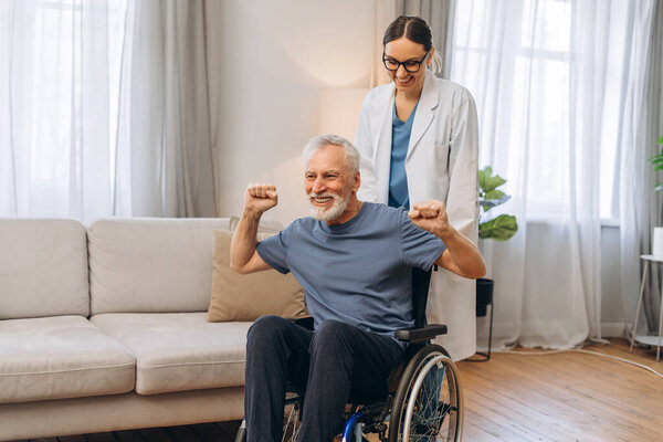 Senior man celebrating recovery with female nurse. Mature patient sitting in wheelchair at home. Rehabilitation concept