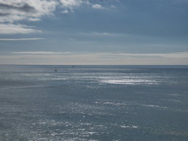 Mavi deniz manzarası, ufukta bulutlar olan sakin dalgalar gökyüzüne dokunuyor. Arka planda tekneler yüzüyor..