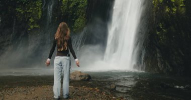 Güçlü şelalenin başında duran turist kız Terjun Munduk, Endonezya, Bali adası. Kadın pozu, arka görüş, yavaş çekim 4K. Güzel doğa manzarası. Seyahat, turizm, tatil