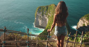 Turist kız dağın tepesinde duran deniz manzarasından hoşlanır. Genç kadın arkası bakış açısı Kelingking Sahili 'ndeki Tyrannosaur kayasının üzerinde dururken rahatlıyor, Nusa Penida, Endonezya. Ünlü turistik yer. Yavaş Hareket 4K