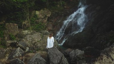 Kadın güçlü şelaleye karşı taş üzerinde meditasyon yapıyor. Beyaz tişörtlü turist kız orman şelalesinin tadını çıkarıyor. Kadın su sıçratarak rahatlıyor. Arka plan, koyu tonlama, İHA uçuşu