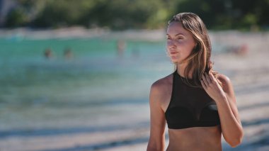Bikini giyen bir kadın kumlu sahilde duruyor ve okyanusa bakıyor. Güneş tepemizde ışıl ışıl parlıyor, sıcak ve manzaralı bir zemin oluşturuyor..