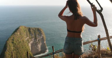 Bir kadın uçurumun tepesinde durmuş, Nusa Penida Adası, Endonezya, Kelingking Gizli Noktası Sahili 'ndeki uçsuz bucaksız okyanusa bakıyor. Uçurumların engebeli güzelliği denizin derin maviliğiyle çelişiyor.