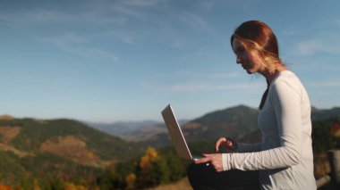 Beyaz kadın, kırsal bir çitin üzerinde oturuyor, dizüstü bilgisayarla yazı yazıyor ve sonbahar günbatımı dağlarının doğasının tadını çıkarıyor. Karpat Dağları 'nda çevrimiçi dijital göçebe işi. Açık hava yaşam tarzı yolculuğu