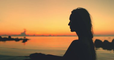 Tayland 'ın turuncu günbatımına karşı çerçevelenmiş kumlu bir sahilde oturan bir kadının silüeti. Durgun sular, ufka doğru bakan renkli gökyüzünü yansıtıyor..