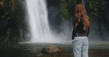 Güçlü bir Bali şelalesinin önünde duran, arka planda çağlayan su olan odaklı bir kadın. Sahne, insan figürü ile doğal mucize arasındaki zıtlığı yakalar..