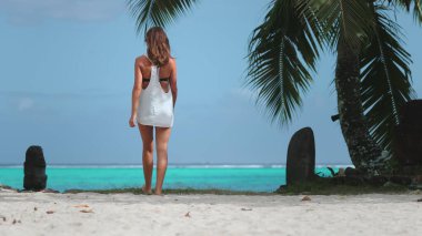 Beyaz elbiseli kadın kumlu sahilde yürüyor, okyanusa bakıyor. Seyahat, turizm, dinlenme konsepti. Palmiye ağacı, turkuaz su. Yaz tatilinde açık hava seyahati. Arka plan
