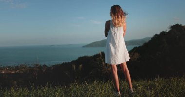 Beyaz elbiseli kadın tepe okyanusunda ve uzakta küçük bir adada duruyor. Kız tropikal adayı rahatlatır, vahşi doğanın tadını çıkarır. Açık hava gezisi, yaz tatili.,