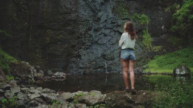 Kadınlar kayaların üzerinde duran orman şelalesinin tadını çıkarırlar. Endonezya 'daki Bali adasına git. Yaz tatilinde açık hava seyahati. Karanlık dramatik tonlama. Arka plan