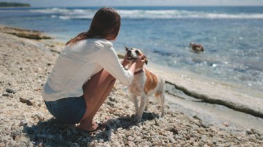 Güneşli bir yaz gününde sahilde köpekle oynayan bir kadın. Genç ve güzel dişi, sevimli hayvanını okşuyor. Neşeli ve kaygısız hissediyorum, plaj gününün tadını çıkarıyorum ve mavi denizde birlikte yürüyoruz.