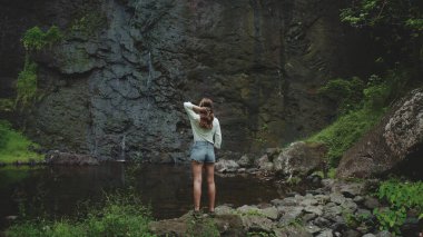 Genç bir kadın gür bir ormanda şelale havuzunun yanında duruyor, uzun saçlarına dokunuyor, Fransız Polinezya Adası 'nın huzurlu manzarasının tadını çıkarıyor.