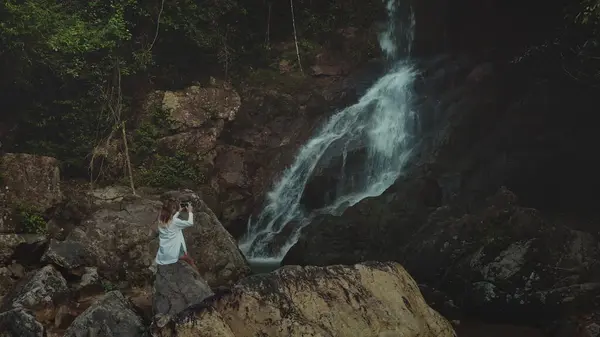 stock image Woman sitting big stone, take picture on mobile phone of waterfall cascade rocky cliff. Female in white shirt relax enjoy beautiful wild nature landscape exotic rainforest. Travel tropical Bali island