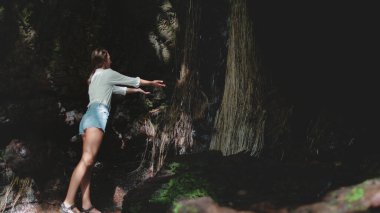 Genç bir kadın, yoğun yaprakların arasından süzülen, karanlık köşelerde gölgeler yaratan, güneş ışığıyla çevrili bir şelaleye dokunmak için uzanan yemyeşil bir mağara keşfeder.