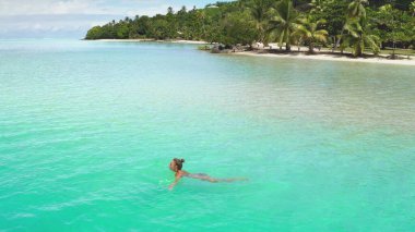 Young woman is swimming in the turquoise lagoon of maupiti island, french polynesia. The image is taken from a drone flying above the water clipart