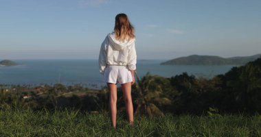 Beyaz elbiseli bir kadın tepe okyanusunda ve uzaktaki küçük bir adada duruyor. Kız tropikal adayı rahatlatır, vahşi doğanın tadını çıkarır. Açık hava gezisi, yaz tatili.,