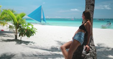 Turist, beyaz plajda güneşli bir günün tadını çıkarıyor. Filipinler, arka planda geleneksel bir yelkenli ile palmiye ağacının altında dinleniyor.