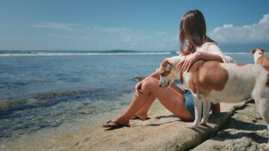 Bir kadın büyük bir kayanın üzerinde küçük bir köpeğin yanında oturuyor. Rahatlamış görünüyor ve önündekilere bakıyor. Köpek onun yanında oturuyor, ona bakıyor..
