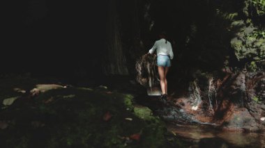 Dişi turist, küçük bir şelalenin arkasındaki bir mağarada kayaların üzerinde dikkatlice yürüyor ve tropikal doğanın güzelliğini keşfediyor.