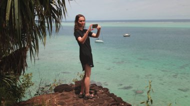 Sahilde bir kadın elinde kamerayla suyun üzerinde süzülen bir teknenin fotoğrafını çekiyor..